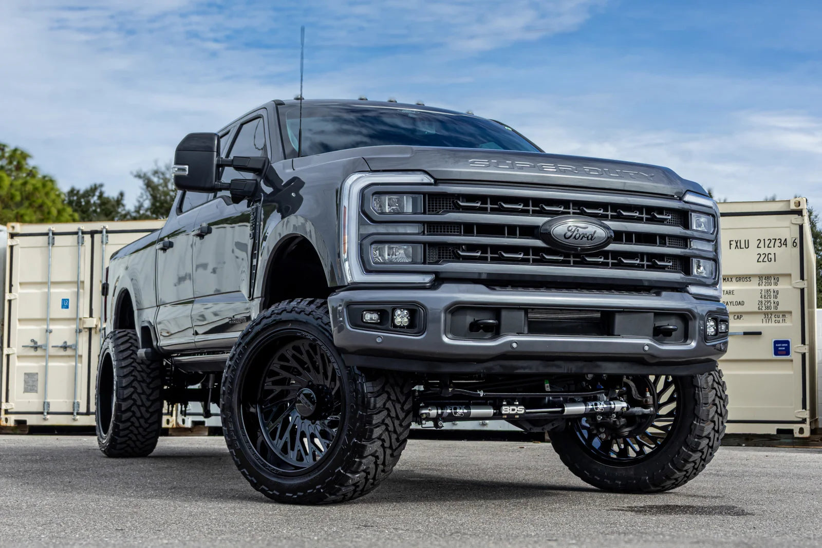 A dark gray lifted Ford pickup truck with oversized black off-road tires and custom wheels, parked outdoors in front of shipping containers under a blue sky.