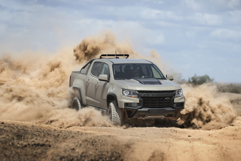A silver Chevrolet Colorado pickup truck driving through dusty desert terrain, kicking up a large cloud of brown sand under a blue sky.